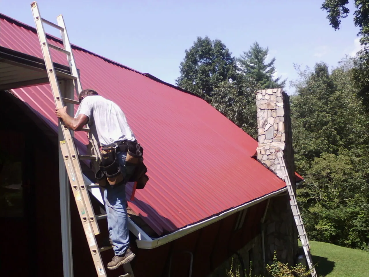 Roofer on a ladder beside a red metal roof and stone chimney — Soffit & Fascia Repair by Roofing Brothers in Valles Mines